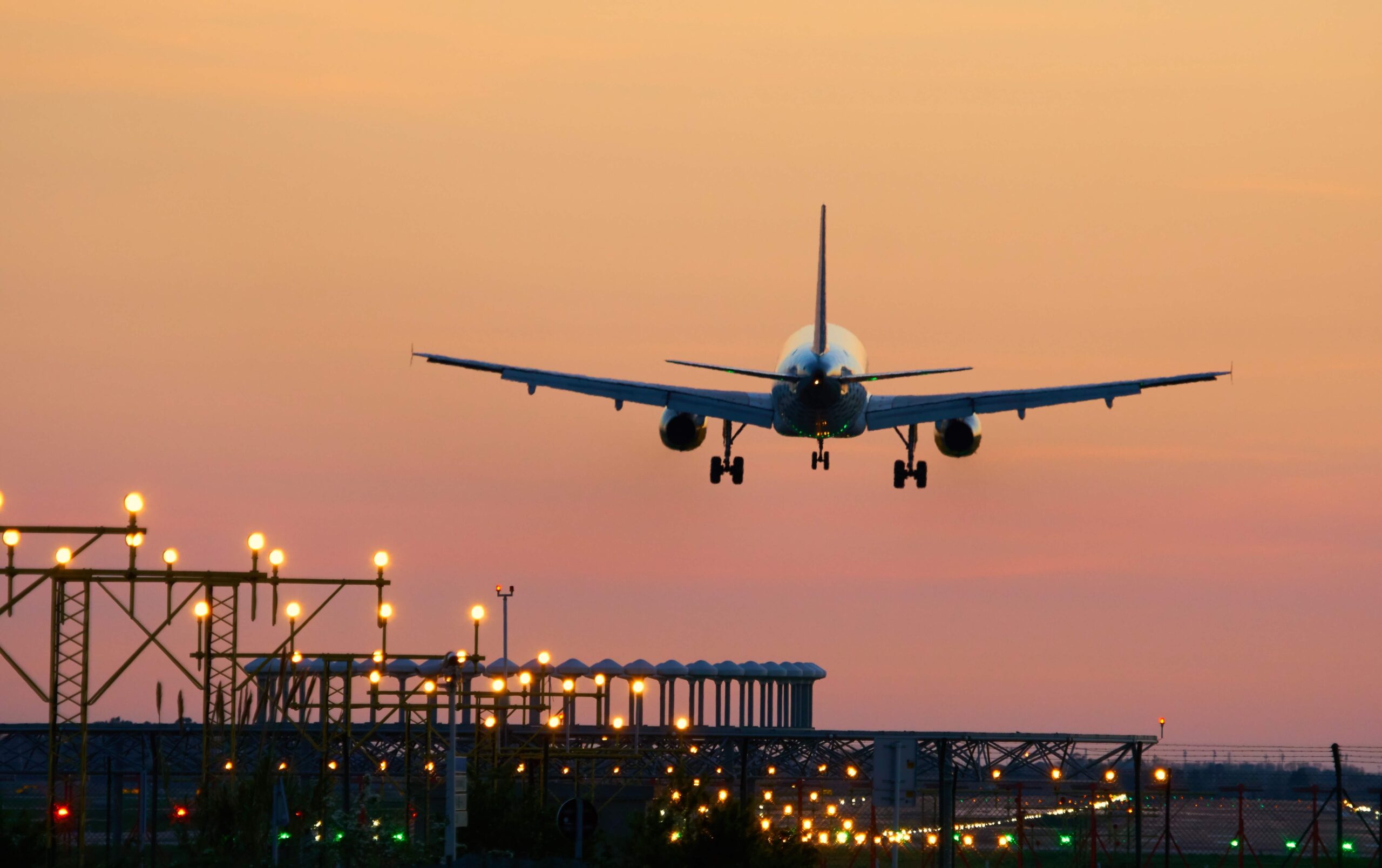 A commercial airplane approaches a runway for landing at sunset, with airport lights illuminating the ground and the sky displaying orange and pink hues.